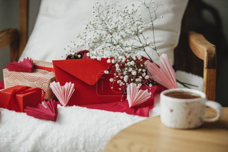 Photo for Happy valentines day! Modern gifts, red envelope and white flowers on cozy armchair on background of defocused stylish cup of tea with heart on wooden table. Valentine morning surprise - Royalty Free Image