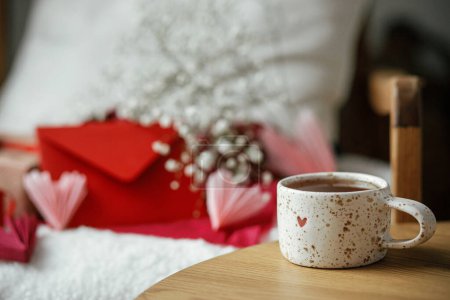 Téléchargez les photos : Cute stylish cup of tea with heart on wooden table against modern armchair with gifts, red envelope and white flowers. Valentine morning surprise for beloved. Happy valentines day! - en image libre de droit