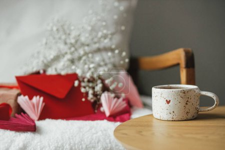 Téléchargez les photos : Happy valentines day! Cute modern cup of tea with heart on wooden table against stylish chair with gifts, red envelope and white flowers. Valentine morning surprise for beloved. Space for text - en image libre de droit