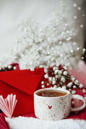 Foto de Happy valentines day! Cute stylish cup of tea with heart on cozy modern armchair with gifts, red envelope and white flowers. Valentine morning surprise for beloved - Imagen libre de derechos