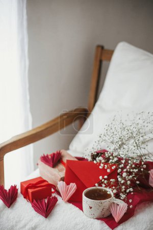Téléchargez les photos : Cute stylish cup of tea with heart on cozy modern armchair with gifts, red envelope and white flowers. Valentine morning surprise for beloved. Happy valentines day! - en image libre de droit