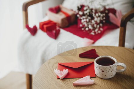 Téléchargez les photos : Happy valentines day! Cute modern cup of tea with heart and red envelope on wooden table against stylish chair with gifts and white flowers. Valentine morning surprise for beloved. Space for text - en image libre de droit