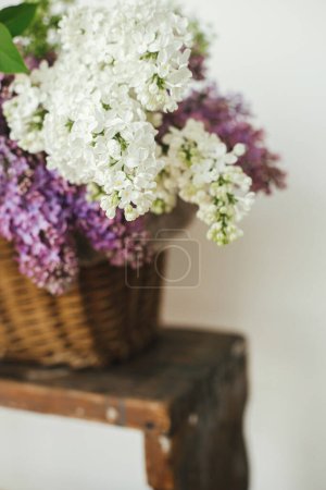 Foto de Beautiful lilac flowers in wicker basket on wooden chair. Purple and white lilacs petals close up, floral composition in home. Spring rustic still life on rural background. Mothers day or wedding - Imagen libre de derechos