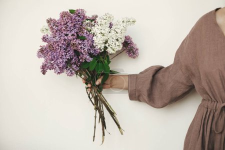Téléchargez les photos : Femme élégante main tenant de belles fleurs lilas contre le mur blanc. Robe femme en lin avec fleurs lilas, vue dégagée. Moment authentique et lunatique. Mariage rustique - en image libre de droit
