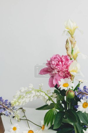Foto de Stylish peony, lupin, iris and daisy arrangement on kenzan with scissors on rustic wood. Creative floral image. Modern summer flowers composition on rustic white table indoors. - Imagen libre de derechos
