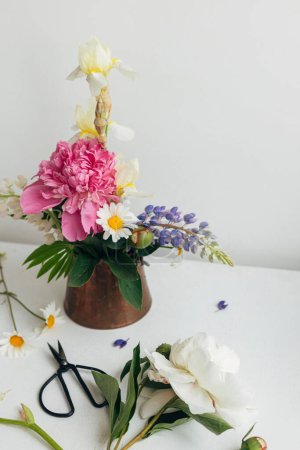 Téléchargez les photos : Modern summer flowers still life arrangement in vintage vase on white wood. Creative floral image. Stylish peony, lupin, iris and daisy composition and scissors on rustic table - en image libre de droit