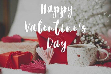 Téléchargez les photos : Happy valentines day text on stylish cup of tea with heart and valentine gifts, red envelope, white flowers. Modern Valentines day greeting card. Handwritten sign - en image libre de droit