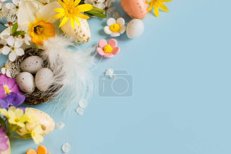 Foto de Happy Easter! Easter eggs in nest, colorful flowers and blooming cherry petals on blue background. Stylish festive template with space for text. Greeting card or banner - Imagen libre de derechos