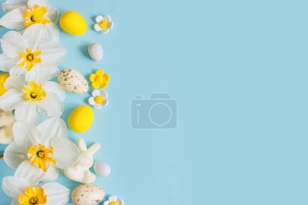Foto de Stylish Easter eggs, bunny and yellow daffodils flowers flat lay on blue background with copy space. Happy Easter! Greeting card template. Modern holiday banner. Festive composition - Imagen libre de derechos