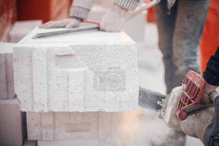Téléchargez les photos : Workers cutting autoclaved aerated concrete block with chainsaw closeup. Builders cutting white blocks for masonry installation. Process of house building at construction site. - en image libre de droit