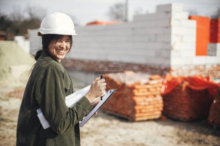 Téléchargez les photos : Young female engineer or construction worker in hardhat smiling against of building new modern house. Stylish happy woman architect with blueprints and plans at construction site - en image libre de droit