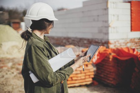 Photo for Stylish woman architect with tablet checking blueprints at construction site. Young female engineer or construction worker in hardhat looking at plans of new modern house - Royalty Free Image