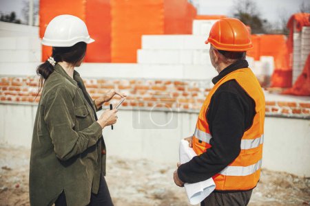 Foto de Stylish woman architect with tablet and senior foreman checking blueprints at construction site. Engineer and construction worker in hardhat looking at plans of new modern house. Teamwork - Imagen libre de derechos