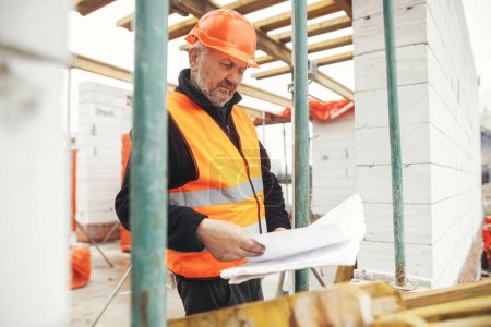 Photo for Senior male foreman in hardhat checking blueprints of building new modern house. Man engineer or construction worker looking at plans at construction site. Copy space - Royalty Free Image