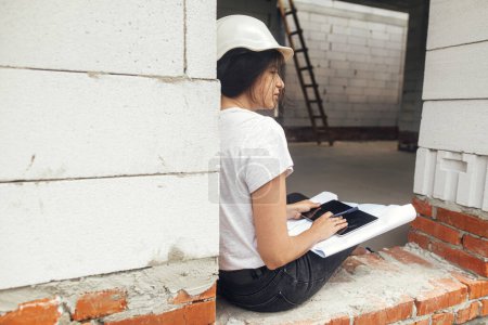 Téléchargez les photos : Young female architect with tablet checking blueprints while sitting in window of new modern house. Stylish woman engineer in hard hat looking at digital plans on tablet at construction site - en image libre de droit
