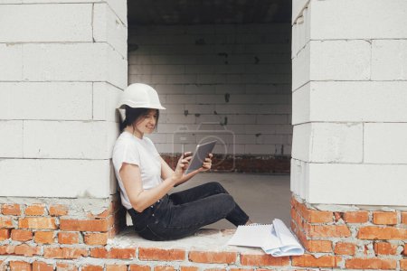 Photo for Stylish happy woman engineer in hard hat looking at digital plans on tablet while sitting in window of new modern house. Young female architect checking blueprints at construction site - Royalty Free Image
