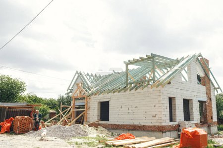Unfinished modern farmhouse building. Wooden roof framing of mansard with dormer and aerated concrete block walls. Timber trusses, rafters and beams on blocks. New house construction