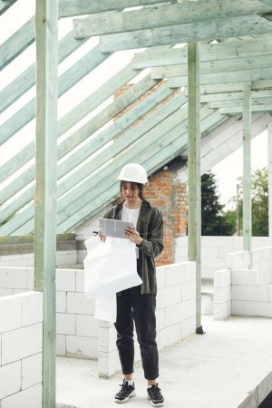 Photo for Stylish woman architect or engineer with tablet checking blueprints against wooden roof framing of modern farmhouse. Young female in hard hat looking at digital plans at construction site - Royalty Free Image