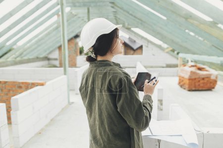 Foto de Stylish woman architect or engineer with tablet checking blueprints against wooden roof framing of modern farmhouse. Young female in hard hat looking at digital plans at construction site - Imagen libre de derechos