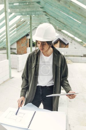 Téléchargez les photos : Young female architect with tablet checking blueprints against wooden roof framing of modern farmhouse. Stylish woman engineer in hard hat looking at digital plans at construction site - en image libre de droit