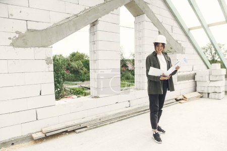 Téléchargez les photos : Stylish happy woman architect checking blueprints against wooden roof framing of modern farmhouse. Young female engineer or construction worker in hardhat with plans at construction site - en image libre de droit