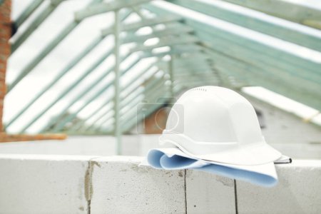 Photo for Architecture and House building concept. Hard hat with house blueprints on aerated concrete blocks on background of unfinished wooden roof framing. Safety helmet on construction site - Royalty Free Image