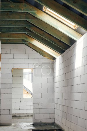 Téléchargez les photos : Unfinished attic wooden roof framing with vapor barrier, dormer and windows. View on timber rafters and beams on aerated concrete blocks in mansard. New modern farmhouse construction - en image libre de droit