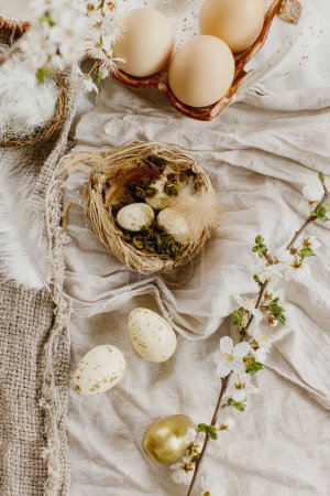 Téléchargez les photos : Happy Easter! Stylish easter eggs and blooming spring flowers on rustic table. Rustic easter still life. Modern eggs in nest and cherry blossoms on linen fabric - en image libre de droit