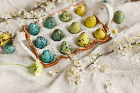 Téléchargez les photos : Happy Easter! Stylish easter eggs and blooming spring flowers on rustic table. Natural painted quail eggs in tray, feathers and cherry blossoms on linen fabric. Rustic easter still life - en image libre de droit