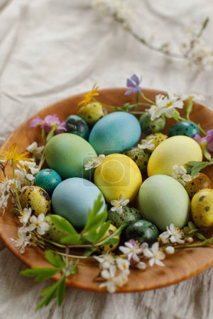 Téléchargez les photos : Happy Easter!  Stylish easter eggs and blooming spring flowers in wooden bowl on rustic table. Natural painted eggs and blossoms on linen fabric. Rustic easter still life - en image libre de droit