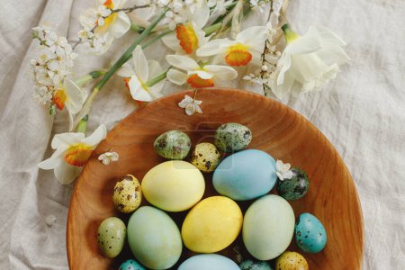 Téléchargez les photos : Rustic easter still life. Stylish easter eggs and blooming daffodils flowers in wooden bowl on linen fabric flat lay. Happy Easter! Natural painted eggs and blossoms on rural table - en image libre de droit