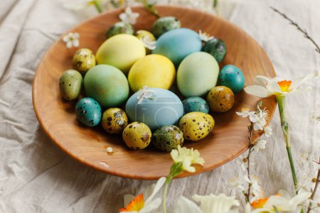 Téléchargez les photos : Joyeux Pâques ! Des œufs de Pâques élégants et des fleurs de printemps fleuries dans un bol en bois sur une table rustique. Oeufs et fleurs peints naturels sur toile de lin. Pâques rustique nature morte - en image libre de droit