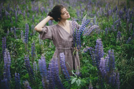 Stylish carefree woman in rustic dress holding lupine bouquet in meadow. Cottagecore aesthetics. Young female in linen dress gathering wildflowers in atmospheric summer countryside, slow life