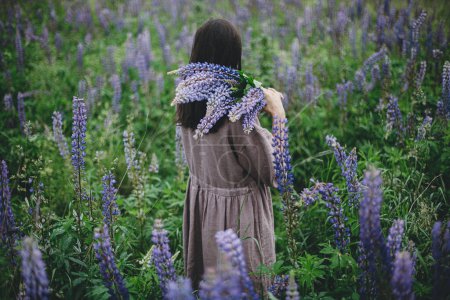 Photo for Stylish woman in rustic dress gathering lupine bouquet in meadow, atmospheric moment. Cottagecore aesthetics. Young female in linen dress holding wildflowers in summer countryside, rural slow life - Royalty Free Image