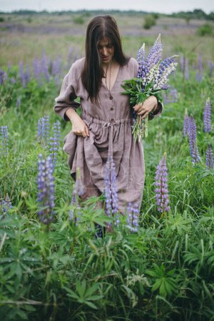 Stilvolle Frau in rustikalem Kleid mit Lupinenstrauß auf der Wiese. Cottagecore-Ästhetik. Junge Frau im Leinenkleid spaziert und sammelt Wildblumen in stimmungsvoller Sommerlandschaft