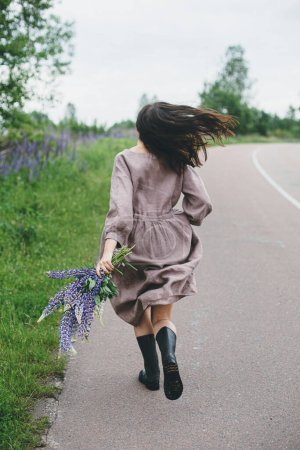 Téléchargez les photos : Femme élégante en robe rustique courant avec bouquet de lupin dans la campagne estivale. Esthétique Cottagecore. Jeune femme en robe de lin relaxant avec des fleurs sauvages après la pluie sur la route rurale - en image libre de droit
