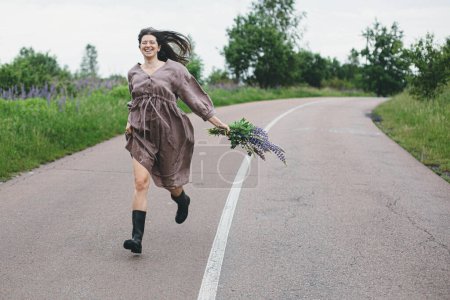 Photo for Happy carefree woman in rustic dress running with lupine bouquet in summer countryside. Cottagecore aesthetics. Young female in linen dress relaxing with wildflowers after rain on rural road - Royalty Free Image