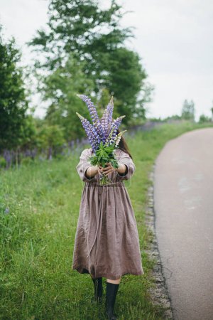Téléchargez les photos : Femme tenant bouquet de lupin fermer dans la campagne d'été. Esthétique Cottagecore. Jeune femme en robe de lin tenant des fleurs sauvages sur fond de route rurale et prairie, vie lente - en image libre de droit
