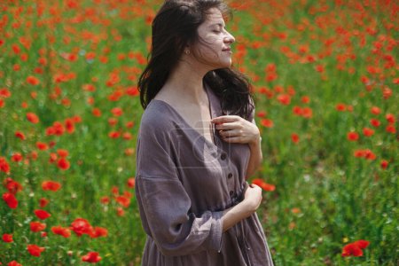 Téléchargez les photos : Stylish sensual woman in rustic dress relaxing in red poppy field. Cottagecore aesthetics. Young female in linen dress enjoying summer among wildflowers meadow in sunny countryside, slow life - en image libre de droit