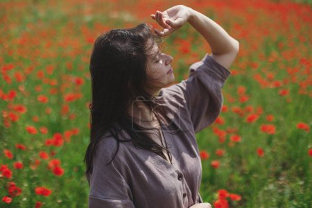 Téléchargez les photos : Stylish sensual woman in rustic dress relaxing in red poppy field. Cottagecore aesthetics. Young female in linen dress enjoying summer among wildflowers meadow in sunny countryside, slow life - en image libre de droit