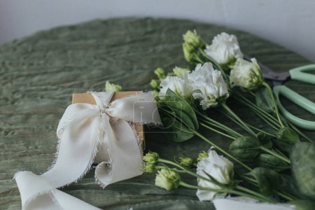 Foto de Stylish simple gift with silk ribbon and beautiful flowers on green fabric. Happy women's day and mother's day. Modern spring holiday still life. Greeting card - Imagen libre de derechos
