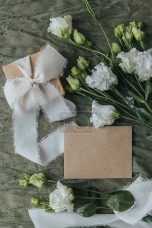 Téléchargez les photos : Stylish empty card, gift and beautiful flowers on green fabric background flat lay. Happy women's day and mother's day concept. Greeting card template, space for text. Eustoma flowers - en image libre de droit