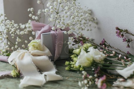 Téléchargez les photos : Happy women's day and mother's day. Stylish simple gift with silk ribbons and beautiful flowers on green fabric against rustic wall. Modern spring holiday still life. Greeting card - en image libre de droit
