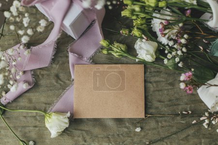 Foto de Stylish empty card, gift and beautiful flowers with ribbons on green fabric background flat lay. Happy women's day and mother's day concept. Greeting card template, space for text - Imagen libre de derechos