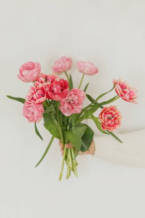 Foto de Elegantes tulipanes rosados en la mano sobre fondo blanco de pared. Mujer sosteniendo hermoso ramo de primavera de cerca, espacio de copia. Saludos con feliz día de las madres y día de las mujeres - Imagen libre de derechos