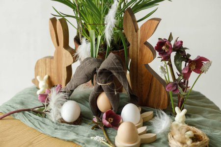 Téléchargez les photos : Joyeux Pâques ! Élégant lapin en bois, fleurs de printemps, œufs naturels et oreilles de lapin serviette sur table rustique dans la chambre. Pâques nature morte. Aménagement festif et décor dans une ferme - en image libre de droit