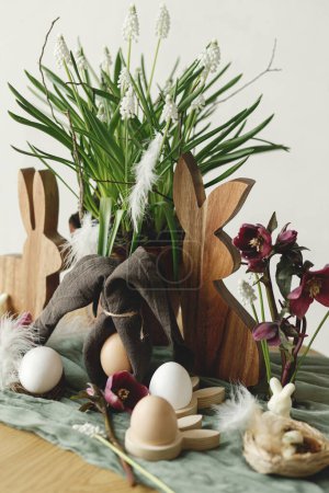 Foto de ¡Feliz Pascua! Elegante conejito de madera, flores de primavera, huevos naturales y orejas de conejito de servilleta en la mesa rústica en la habitación. Naturaleza muerta de Pascua. Disposición festiva y decoración en casa de campo - Imagen libre de derechos