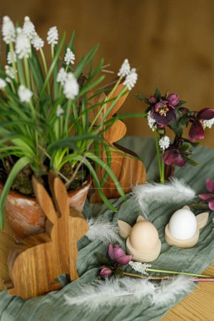 Foto de ¡Feliz Pascua! Elegante conejito de madera, flores de primavera, huevos naturales y plumas en la mesa rústica en la habitación. Naturaleza muerta de Pascua. Disposición festiva y decoración en casa de campo - Imagen libre de derechos