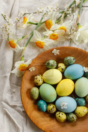 Téléchargez les photos : Joyeux Pâques ! Pose plate rustique de Pâques. Des œufs de Pâques élégants et des fleurs de printemps fleuries dans un bol en bois sur une table rustique. Œufs et jonquilles peints naturels sur toile de lin - en image libre de droit