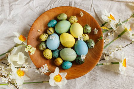 Foto de Naturaleza muerta de Pascua rústica. Elegantes huevos de Pascua y flores florecientes de primavera en un tazón de madera sobre tela de lino puesta plana. ¡Feliz Pascua! Huevos naturales pintados y flores en la mesa rural - Imagen libre de derechos